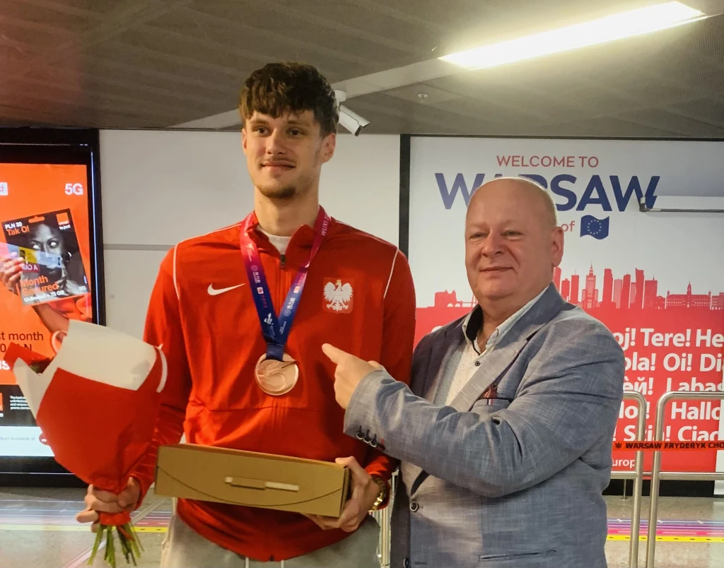 Młody mężczyzna w sportowej czerwonej bluzie z herbem Polski na piersi trzyma kwiaty i pudełko, na szyi zawieszony brązowy medal. Obok niego stoi starszy mężczyzna w jasnej marynarce wskazujący na młodego sportowca z uśmiechem. W tle baner z napisem Wa...