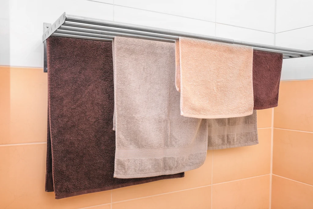 Mokre ręczniki zaleca się suszyć poza łazienką Trzy ręczniki w różnych odcieniach beżu i brązu wiszą na metalowym wieszaku w łazience z jasno-pomarańczowymi kafelkami na ścianie.