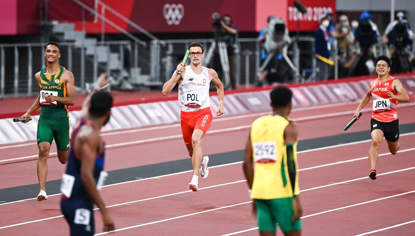 Biegacze z czterech różnych reprezentacji narodowych uczestniczą w sztafecie na stadionie lekkoatletycznym podczas igrzysk olimpijskich. W tle widać trybuny i kilku fotoreporterów. Każdy zawodnik trzyma pałeczkę sztafetową, zawodnik z Polski znajduje s...