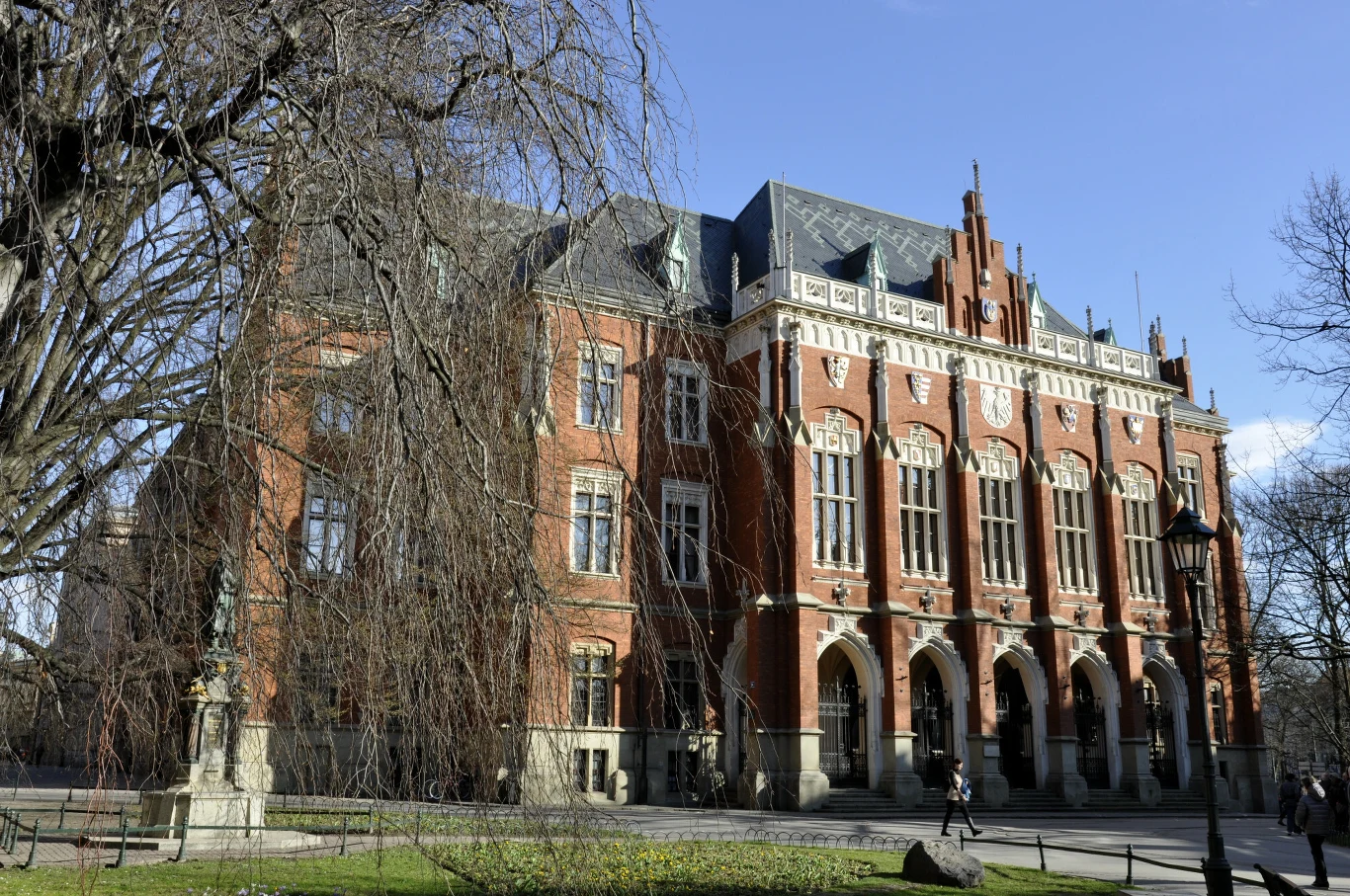 Na którym miejscu uplasowała się Polska? Zabytkowy gmach z czerwonej cegły z licznymi dużymi oknami i ozdobnymi szczytami, otoczony drzewami i zielenią w parkowej przestrzeni, przy którym widoczne są postacie przechodzących osób.