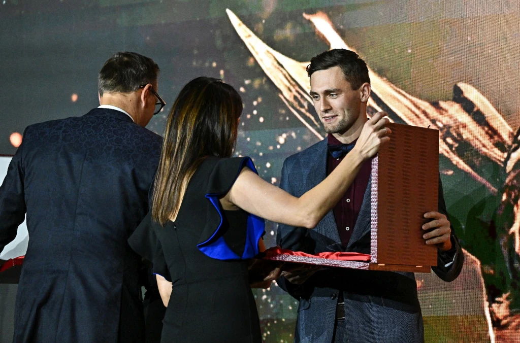 Dariusz Kowaluk Trzech elegancko ubranych ludzi bierze udział w ceremonii wręczenia nagrody, kobieta wręcza pudełko mężczyźnie stojącemu na wprost niej, w tle wyświetlony jest graficzny motyw skrzydeł.