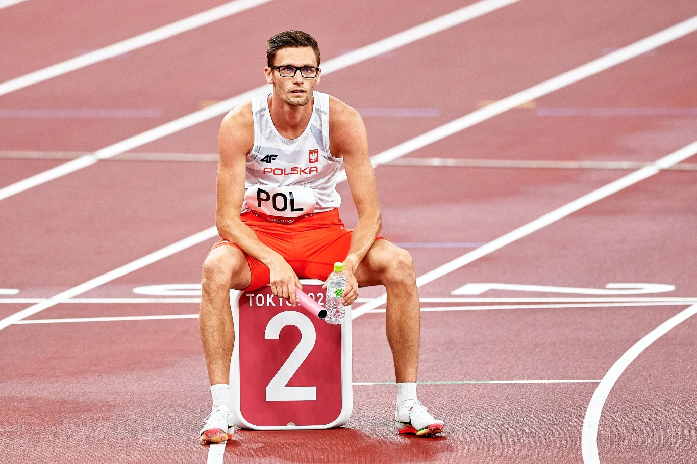 Mężczyzna w stroju reprezentacji Polski siedzi na oznaczeniu toru z numerem 2 na stadionie lekkoatletycznym, trzymając wodę w ręku. W tle widoczne linie bieżni.