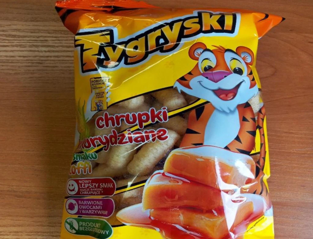 Kolorowe opakowanie chrupków kukurydzianych o smaku toffi z wizerunkiem uśmiechniętego tygrysa i grafiką przedstawiającą karmelowy syrop. Na opakowaniu znajdują się informacje o nowym smaku i naturalnych składnikach.