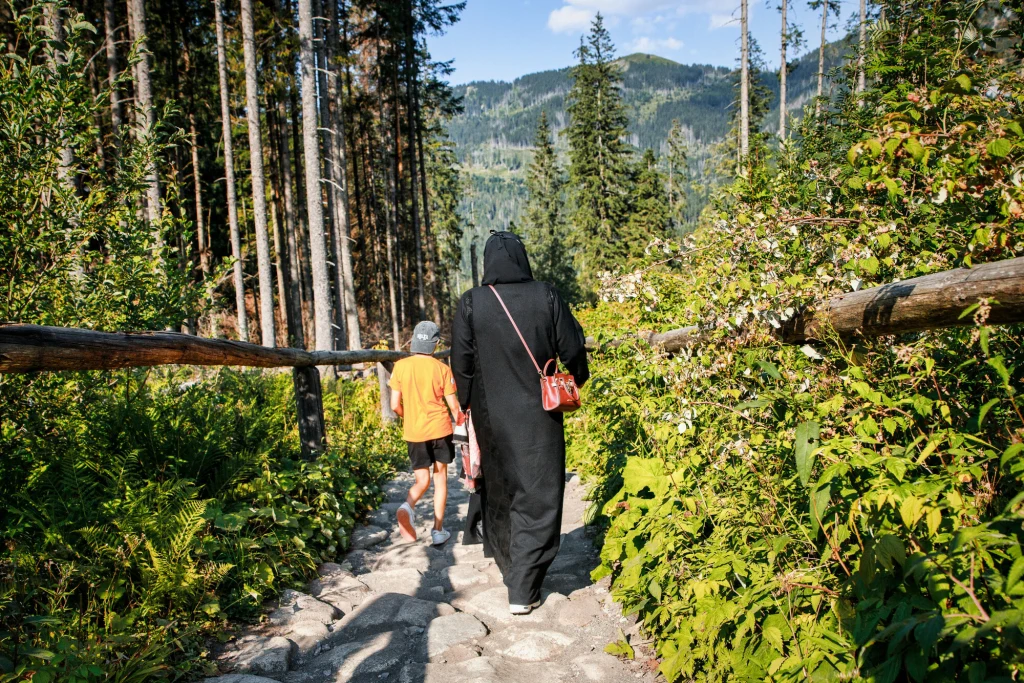 Arabscy turyści są zachwyceni naturą Dwie osoby, dorosła ubrana w czarną abaję oraz dziecko w żółtej koszulce, idą kamienistą ścieżką wśród lasu, otoczonej drewnianym ogrodzeniem, w tle widać górskie szczyty i niebo.