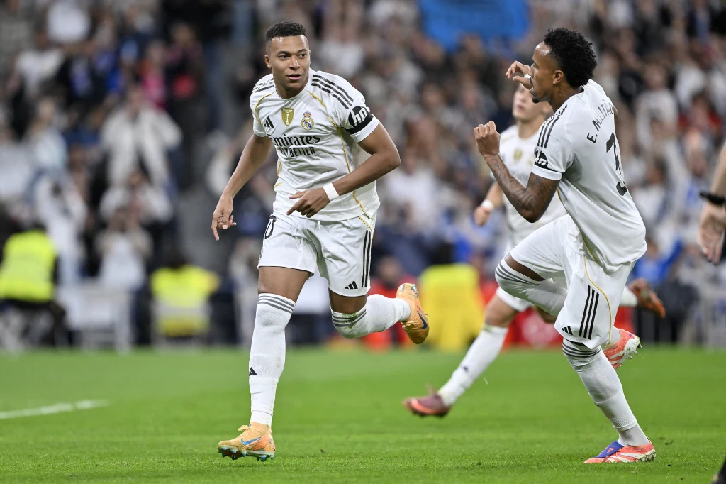 Kylian Mbappe i Eder Militao w barwach Realu Madryt Dwóch piłkarzy w białych strojach Realu Madryt biegnie po murawie stadionu podczas meczu, jeden z nich wskazuje palcem w kierunku drugiego, a w tle widoczni są kibice.