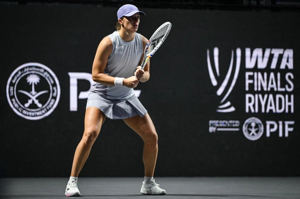Tenisistka przygotowująca się do odbioru piłki podczas meczu, ujęta w sportowej pozycji na tle czarnych, podświetlonych tablic z logotypami turnieju WTA Finals Riyadh.