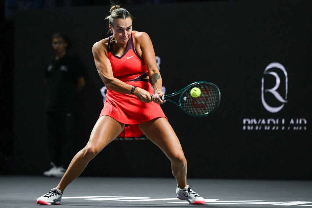 Aryna Sabalenka podczas WTA Finals 2025 Tenisistka w czerwonej sportowej sukience uderza piłkę rakietą podczas meczu tenisowego na czarnym tle z widocznym logo sponsora.