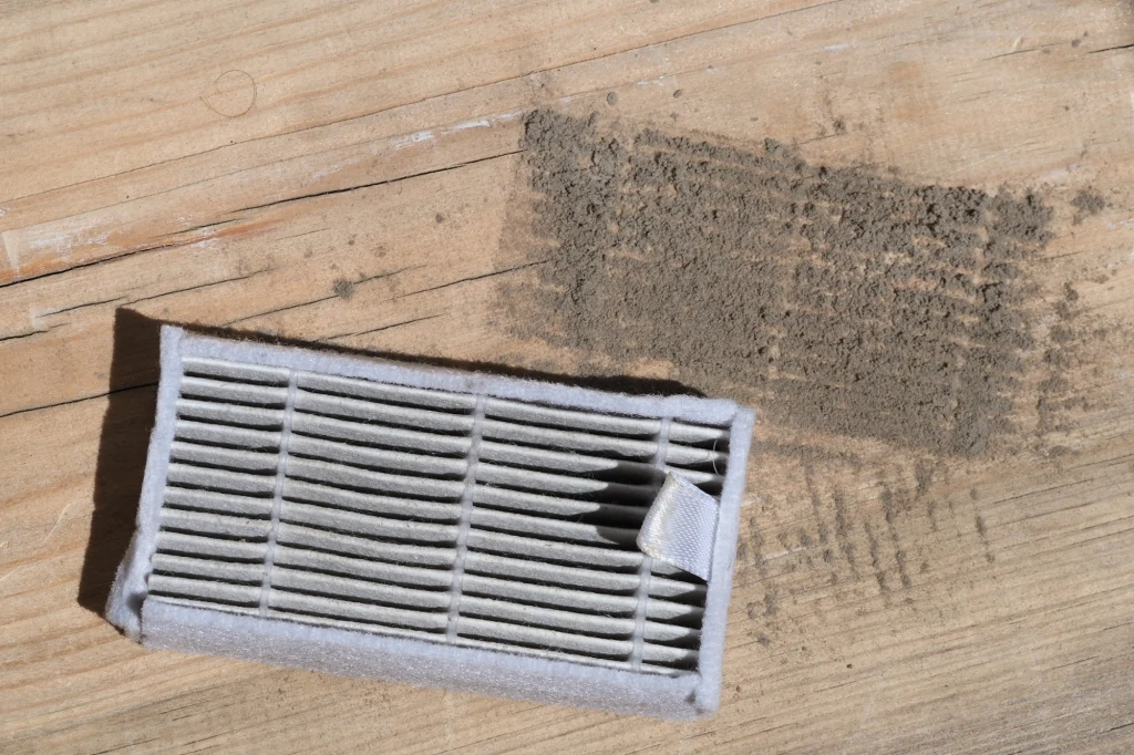 Zużyty filtr powietrza leży na drewnianej powierzchni obok rozsypanego zabrudzenia, prawdopodobnie zebranego przez filtr w trakcie użytkowania.