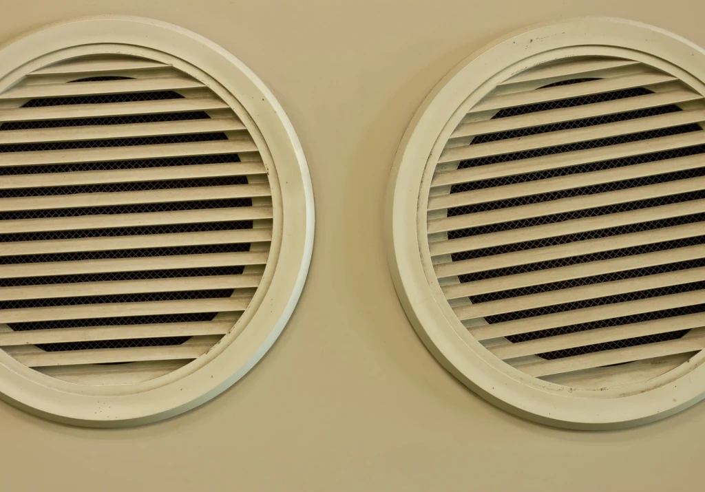 Dwie okrągłe, białe kratki wentylacyjne umieszczone na jasnej ścianie, z ukośnymi żaluzjami blokującymi widok wnętrza.