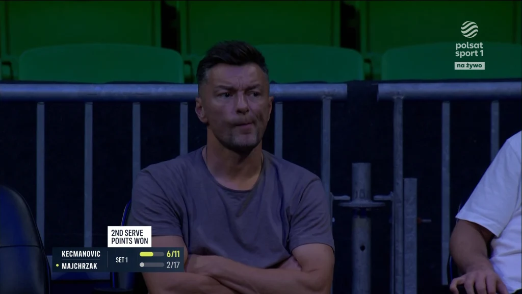 Mężczyzna w szarej koszulce siedzący wśród zielonych krzeseł na trybunach podczas meczu tenisowego, skupiony i patrzący na wydarzenia na korcie, po lewej stronie widoczne informacje o statystykach meczu.