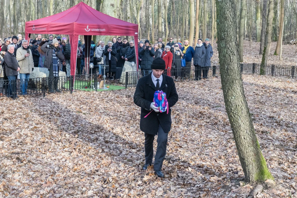 Mężczyzna w garniturze i czarnym płaszczu niesie kolorową urnę, zbliżając się do miejsca pochówku w lesie, gdzie zgromadziła się duża grupa osób pod czerwonym namiotem podczas ceremonii pożegnalnej.