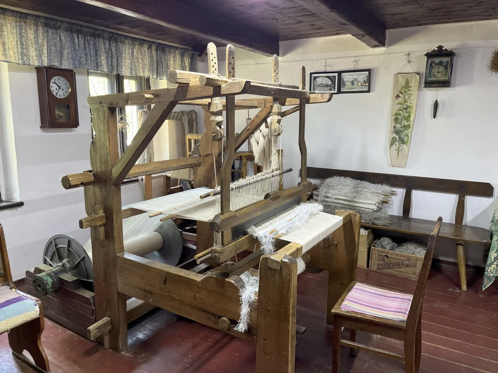 W jednym z domów możemy podziwiać zabytkowe kołowrotki, krosna, żelazka i przedmioty związane z tkactwem Drewniany warsztat tkacki z rozpiętą białą tkaniną ustawiony w tradycyjnym wnętrzu ze starymi meblami, krzesłem, ławą oraz dekoracjami ściennymi, w tle zegar i wiklinowy kosz.
