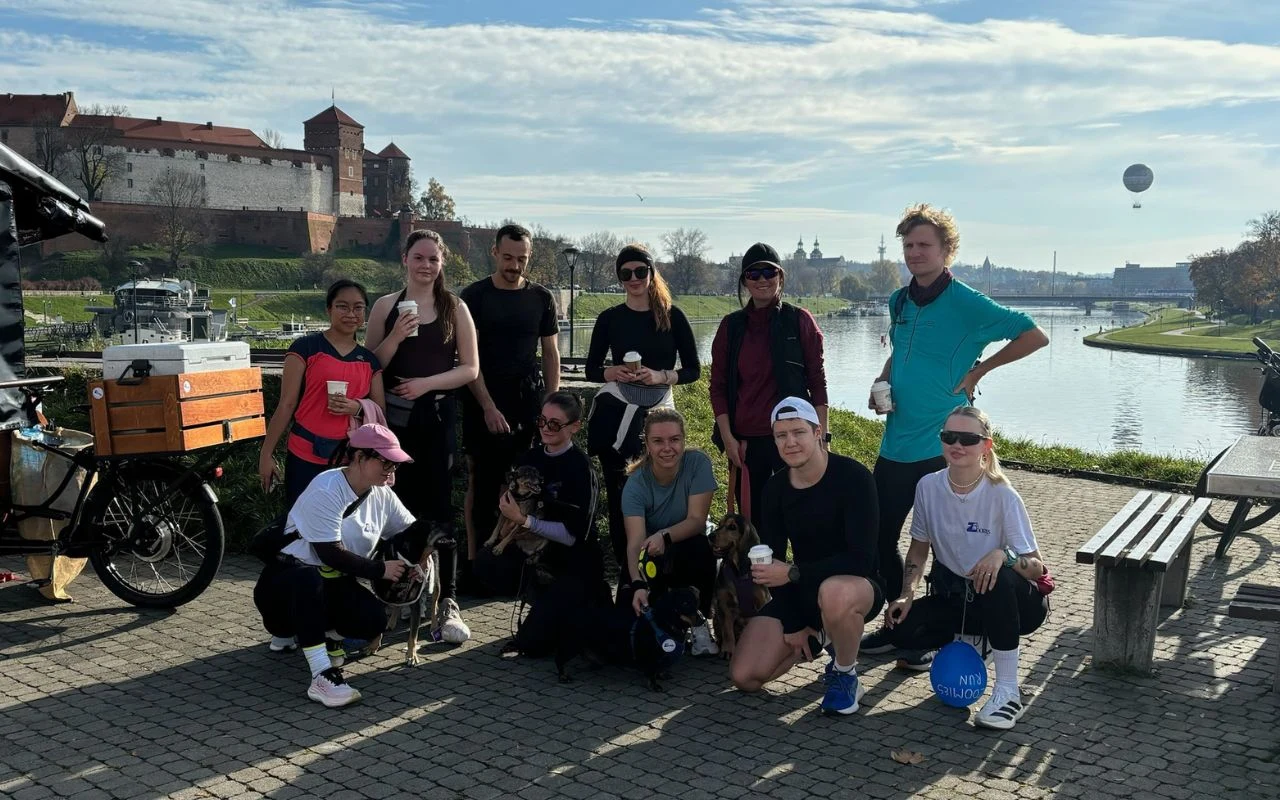 Grupa młodych ludzi ubranych w sportowe stroje pozuje razem na deptaku przy rzece. W tle widać fragment zamku, kosze na rowery i wózek gastronomiczny. Część osób trzyma napoje, a atmosfera wskazuje na aktywność rekreacyjną lub towarzyską na świeżym pow...