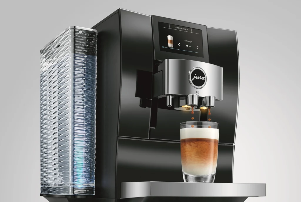Ekspres JURA Z10. Źródło: Oficjalne zdjęcie producenta Nowoczesny ekspres do kawy marki Jura parzy kawę typu latte macchiato. Szkło na tackce prezentuje estetyczną warstwową kawę, a po lewej stronie widoczny jest elegancki, żebrowany pojemnik na wodę.