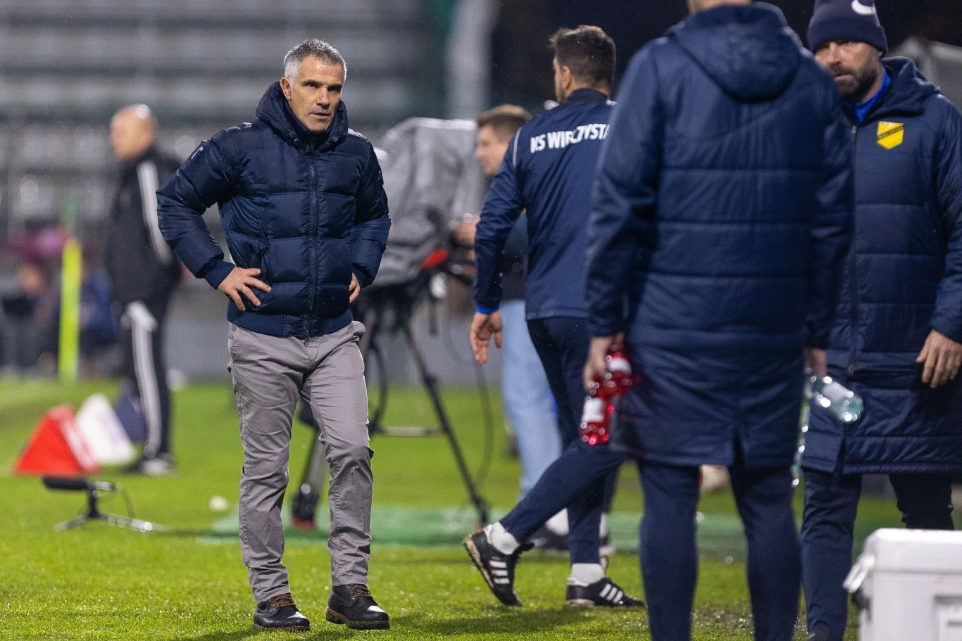 Gino Lettieri Mężczyzna w sportowej kurtce stoi na murawie stadionu, wokół niego kilka innych osób ubranych w drużynowe stroje, tło rozmyte, widać fragmenty sprzętu sportowego i trybuny.