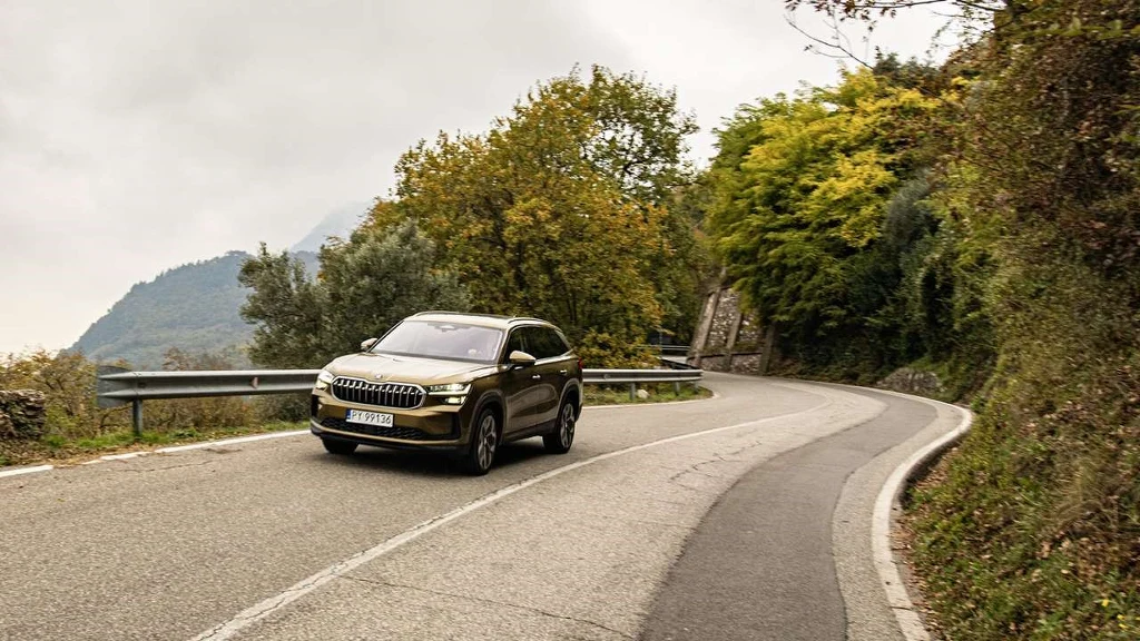 Nowoczesny samochód osobowy w kolorze brązowym jedzie po krętej, asfaltowej drodze otoczonej zielenią i jesiennymi drzewami, w tle widoczne są wzgórza oraz pochmurne niebo.