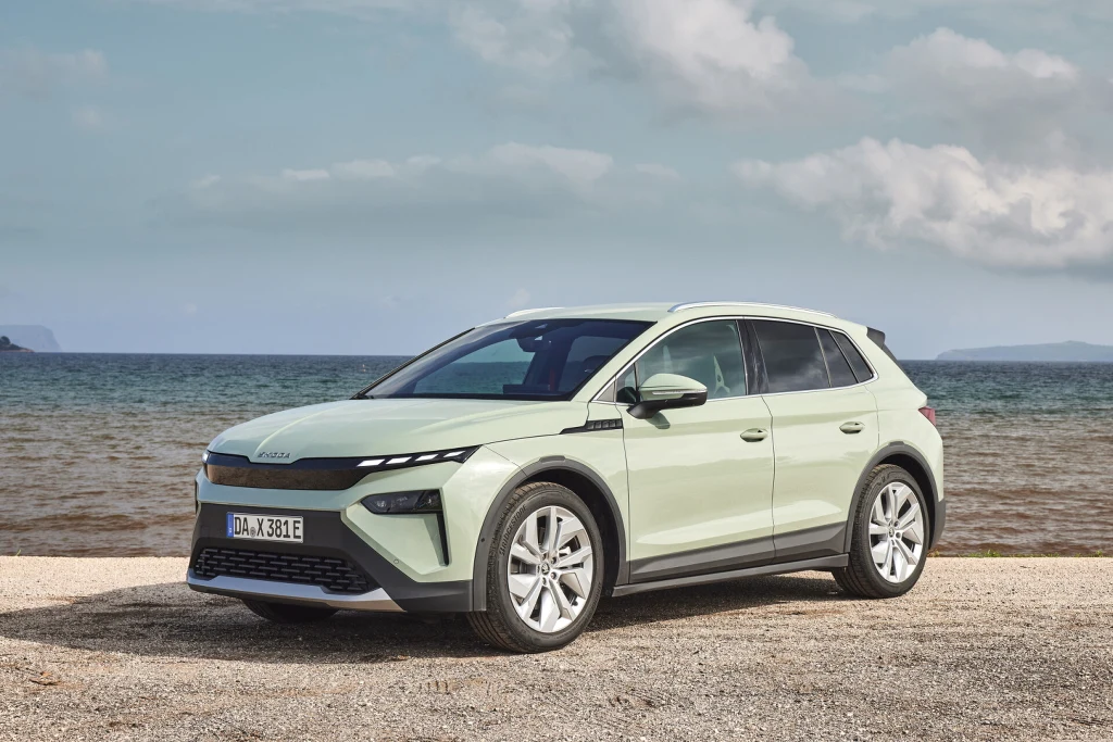 Jasnozielony samochód elektryczny SUV stoi zaparkowany na żwirowej nawierzchni przy brzegu morza, w tle widoczne spokojne fale oraz błękitne niebo z kilkoma chmurami.