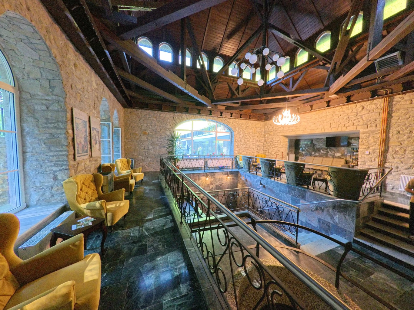 Wnętrze eleganckiego budynku z kamiennymi ścianami, siedzeniami z miękkimi, żółtymi fotelami, dużymi oknami, otwartą antresolą, nowoczesnym oświetleniem oraz efektownymi żyrandolami pod drewnianym stropem.