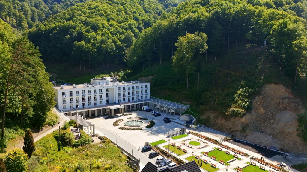 Hotel o białej elewacji położony u stóp zalesionego wzgórza, otoczony dobrze utrzymanymi ogrodami, fontanną oraz tarasami z miejscami do siedzenia, na pierwszym planie ogród i parking.