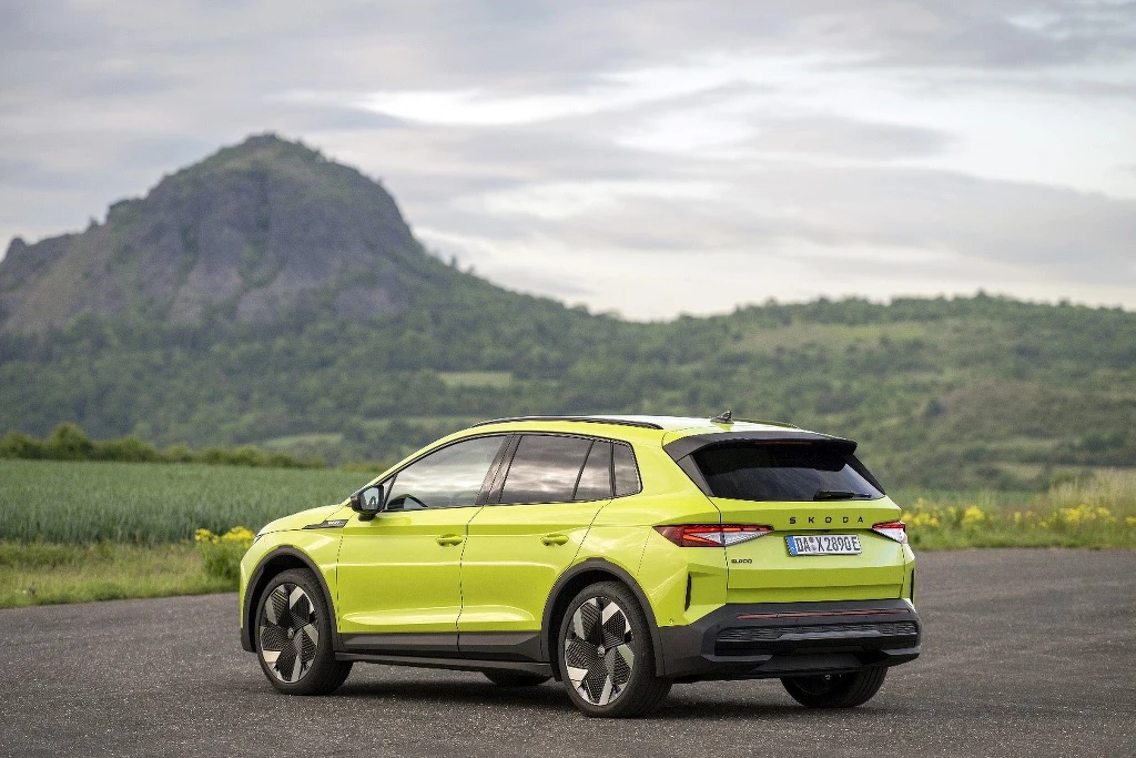 Skoda Elroq Jasnozielony samochód Skoda Enyaq zaparkowany na nawierzchni asfaltowej na tle zielonych pól i wzgórza porośniętego roślinnością pod pochmurnym niebem.