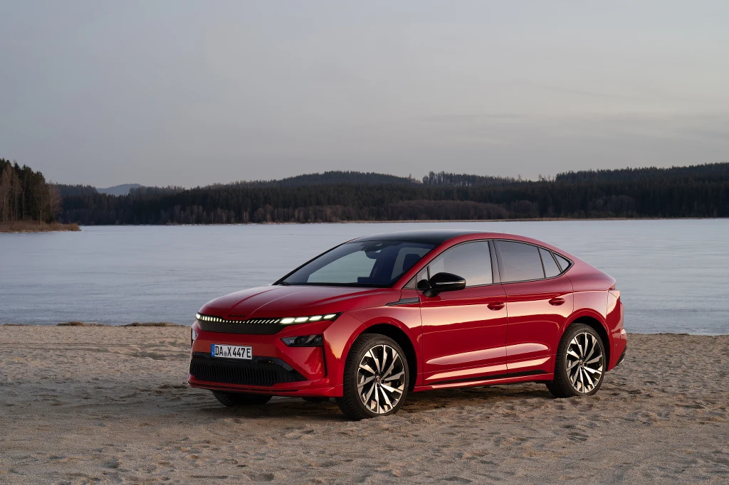 Skoda Enyaq Coupe 85x Czerwony samochód elektryczny zaparkowany na piasku nad brzegiem jeziora, w tle spokojna woda oraz drzewa na horyzoncie. Jasne niebo i łagodne światło tworzą spokojną atmosferę.