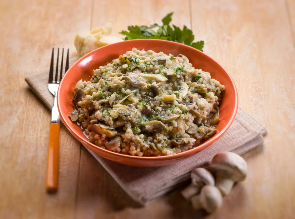 Risotto najlepiej smakuje posypane parmezanem i natką pietruszki Kremowe risotto z pieczarkami, posypane świeżą natką pietruszki, podane na pomarańczowym talerzu, obok sztućce, świeże pieczarki oraz ser na drewnianym stole.