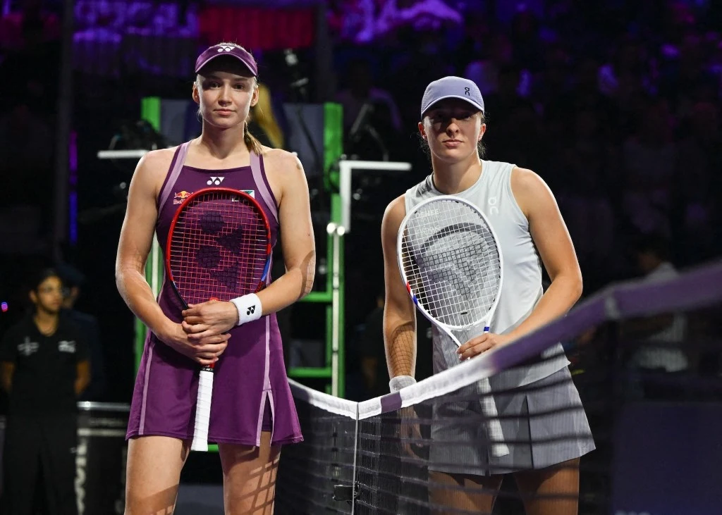 Dwie tenisistki stoją po przeciwnych stronach siatki na korcie, trzymając rakiety i ubrane w sportowe stroje. W tle niewyraźni obserwatorzy oraz sędzia na podwyższeniu.