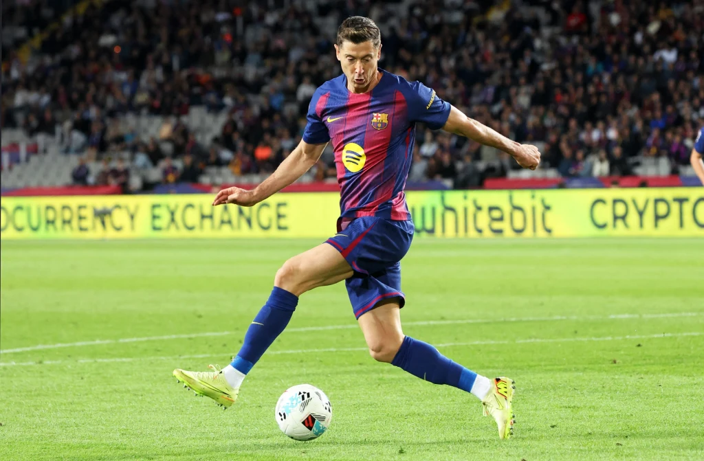 Piłkarz w stroju FC Barcelony wykonuje dynamiczne kopnięcie piłki na boisku podczas meczu, w tle widoczna trybuna pełna kibiców oraz reklamy na bandach.