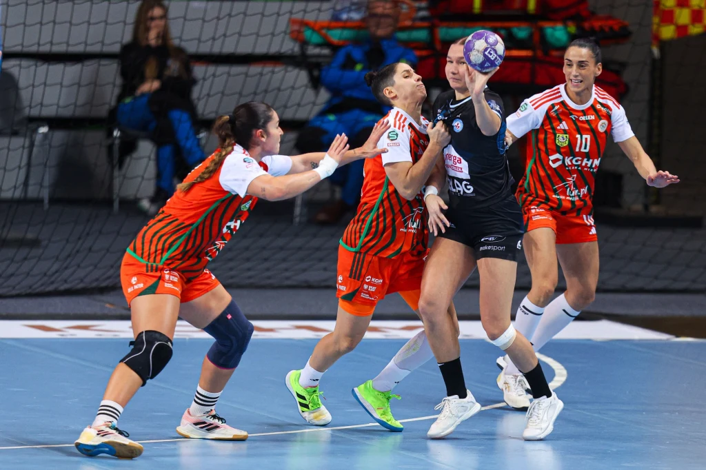 Trzy zawodniczki drużyny w pomarańczowych strojach próbują zatrzymać zawodniczkę w czarnym stroju podczas meczu piłki ręcznej w hali sportowej. W tle widać trybuny i siatkę bramki.