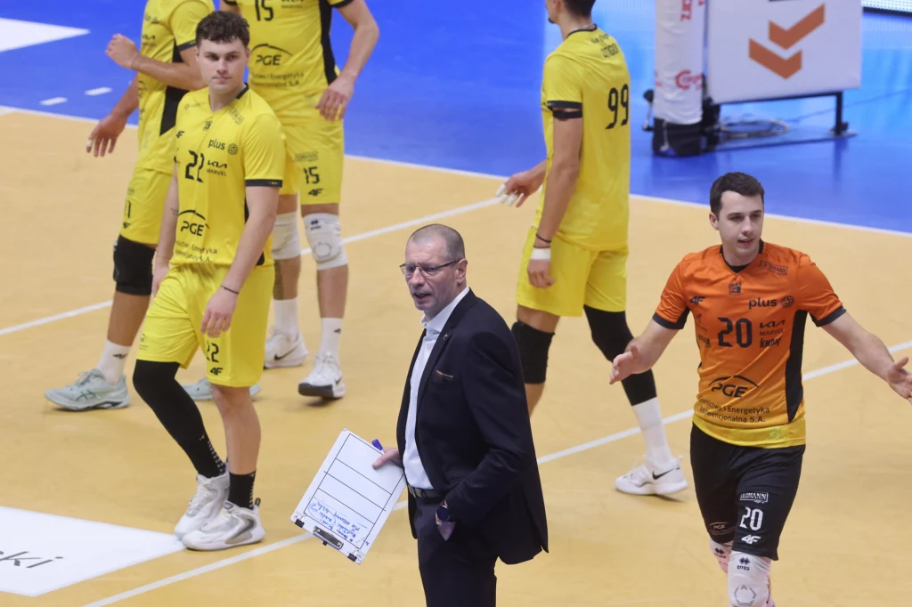 Siatkarze i trener PGE GiEK Skry Bełchatów Grupa siatkarzy ubranych w żółte i pomarańczowe stroje sportowe na hali widowiskowo-sportowej, w centrum uwagi trener z taktyczną tablicą, wyraźna atmosfera meczu i koncentracja zawodników.