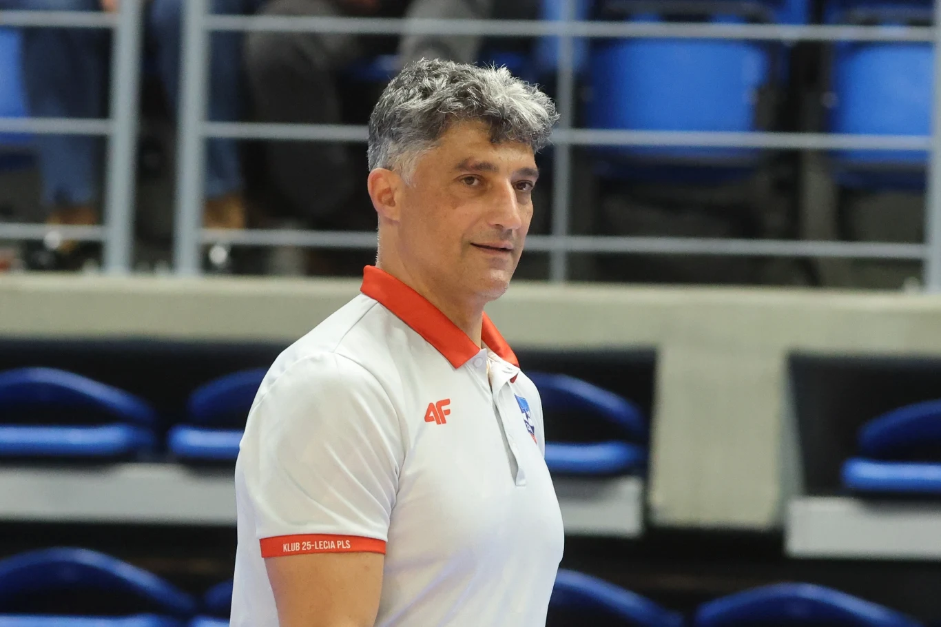 Mężczyzna ubrany w białą koszulkę polo z czerwonym kołnierzem stoi na tle niebieskich siedzeń w hali sportowej. Ujęcie skupia się na jego postaci, przedstawiając go w półprofilu.