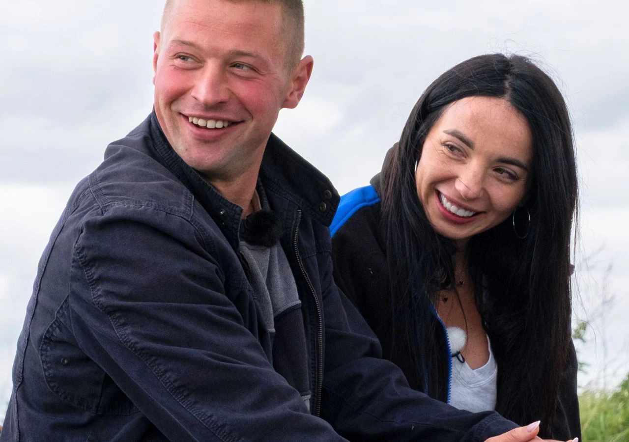 Krzysztof i Agnieszka w "Rolnik szuka żony" Dwoje młodych ludzi siedzi blisko siebie na świeżym powietrzu, oboje uśmiechają się szeroko i patrzą w różne strony. Kobieta ma długie, ciemne włosy, a mężczyzna jest krótko ostrzyżony i ubrany w ciemną kurtkę. Oboje sprawiają wrażenie zadowolonych.