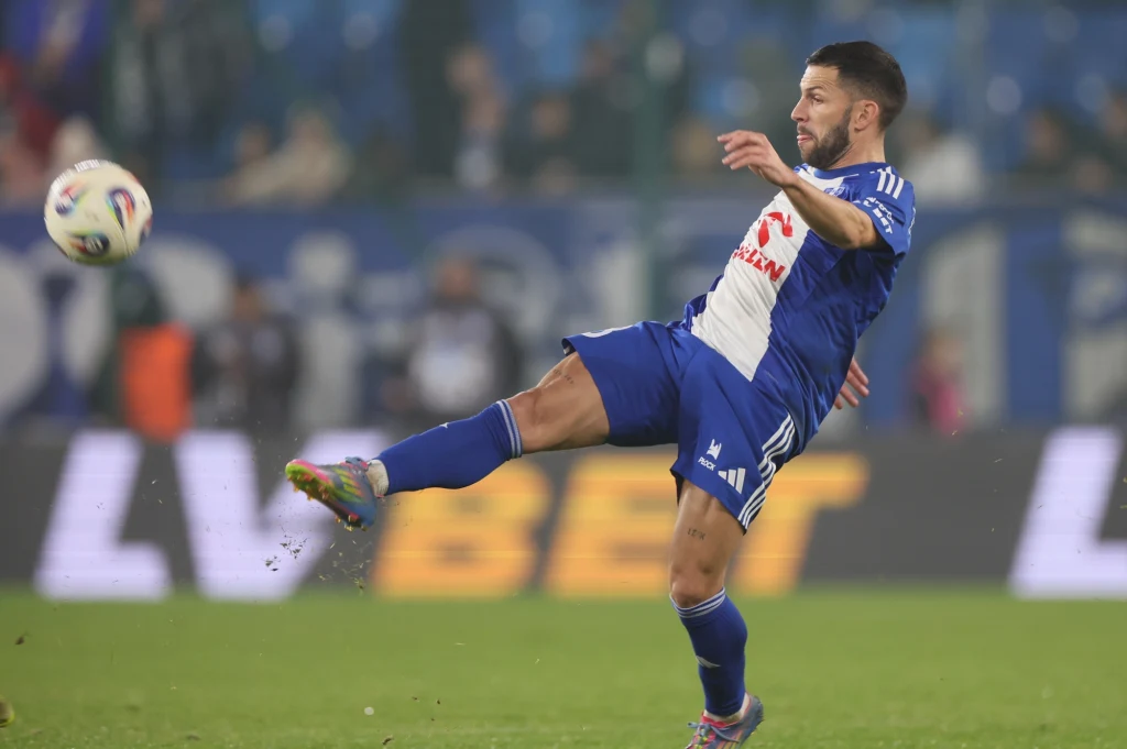 Dani Pacheco, bohater poniedziałkowego wieczoru Piłkarz w niebiesko-białym stroju kopie piłkę podczas meczu piłki nożnej, dynamiczna akcja na zielonej murawie, w tle widoczni kibice oraz reklamy przy boisku.