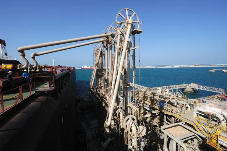 Instalacja do produkcji skroplonego gazu położona na północ od Dohy Zaawansowana infrastruktura portowa z rozbudowanymi konstrukcjami metalowymi oraz systemem rur wykorzystywanym do przeładunku surowców z tankowców. W tle turkusowe morze oraz daleka linia nabrzeża.