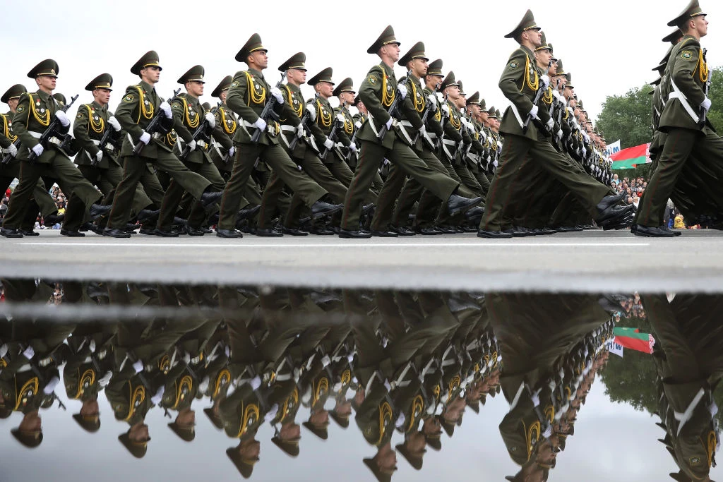 Białoruś deklaruje gotowość wysłania na Ukrainę sił pokojowych Oddział żołnierzy w zielonych mundurach maszeruje synchronicznie po asfalcie podczas defilady wojskowej, ich sylwetki odbijają się w kałuży na pierwszym planie, w tle widoczny fragment flagi.