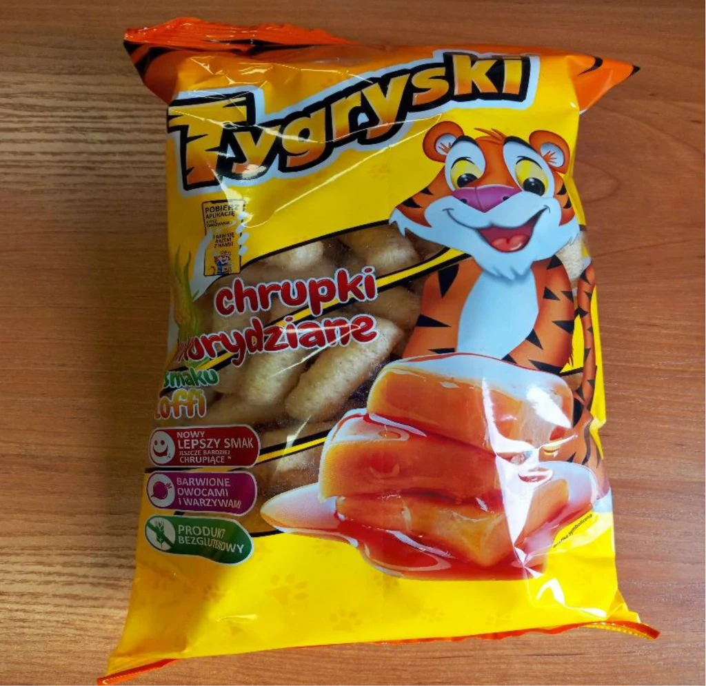 Kolorowe opakowanie chrupek kukurydzianych marki Tygryski z motywem uśmiechniętego tygrysa, grafiką kukurydzianych chrupek oraz miodu, umieszczone na drewnianym tle.