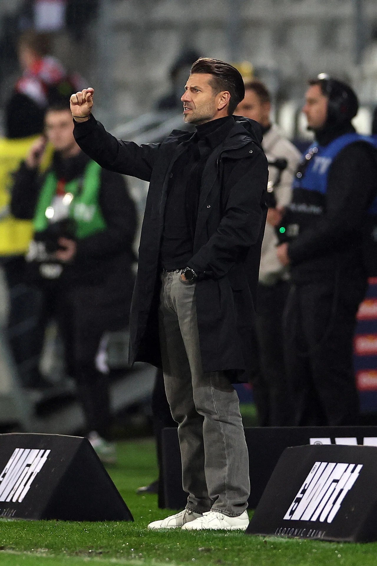 Mężczyzna w ciemnym płaszczu stoi przy linii bocznej boiska, wykonując gest ręką, w tle widoczni są inni ludzie oraz elementy infrastruktury stadionowej.