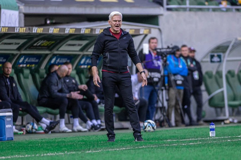 Jacek Magiera na liście kandydatów na trenera Legii Warszawa Trener stojący przy linii bocznej boiska, wydający polecenia, w tle ławka rezerwowych z siedzącymi zawodnikami i sztabem, oraz ekipa kamerzystów rejestrujących wydarzenia sportowe.