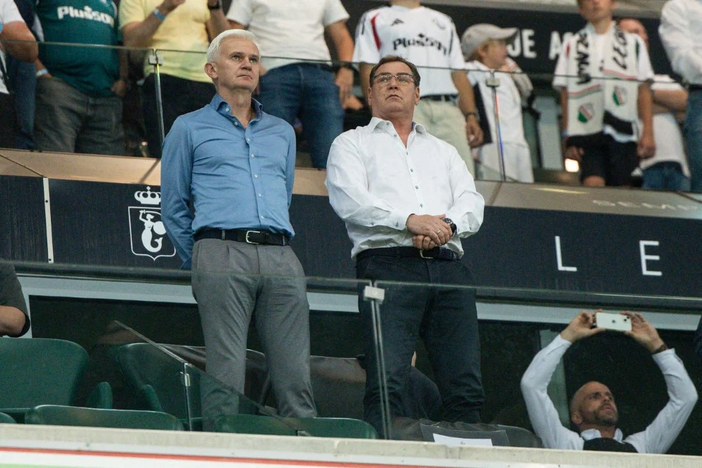 Dwóch mężczyzn w eleganckich koszulach stoi na trybunach stadionu, koncentrując wzrok na wydarzeniach na boisku, w tle inni kibice oraz elementy infrastruktury stadionu.