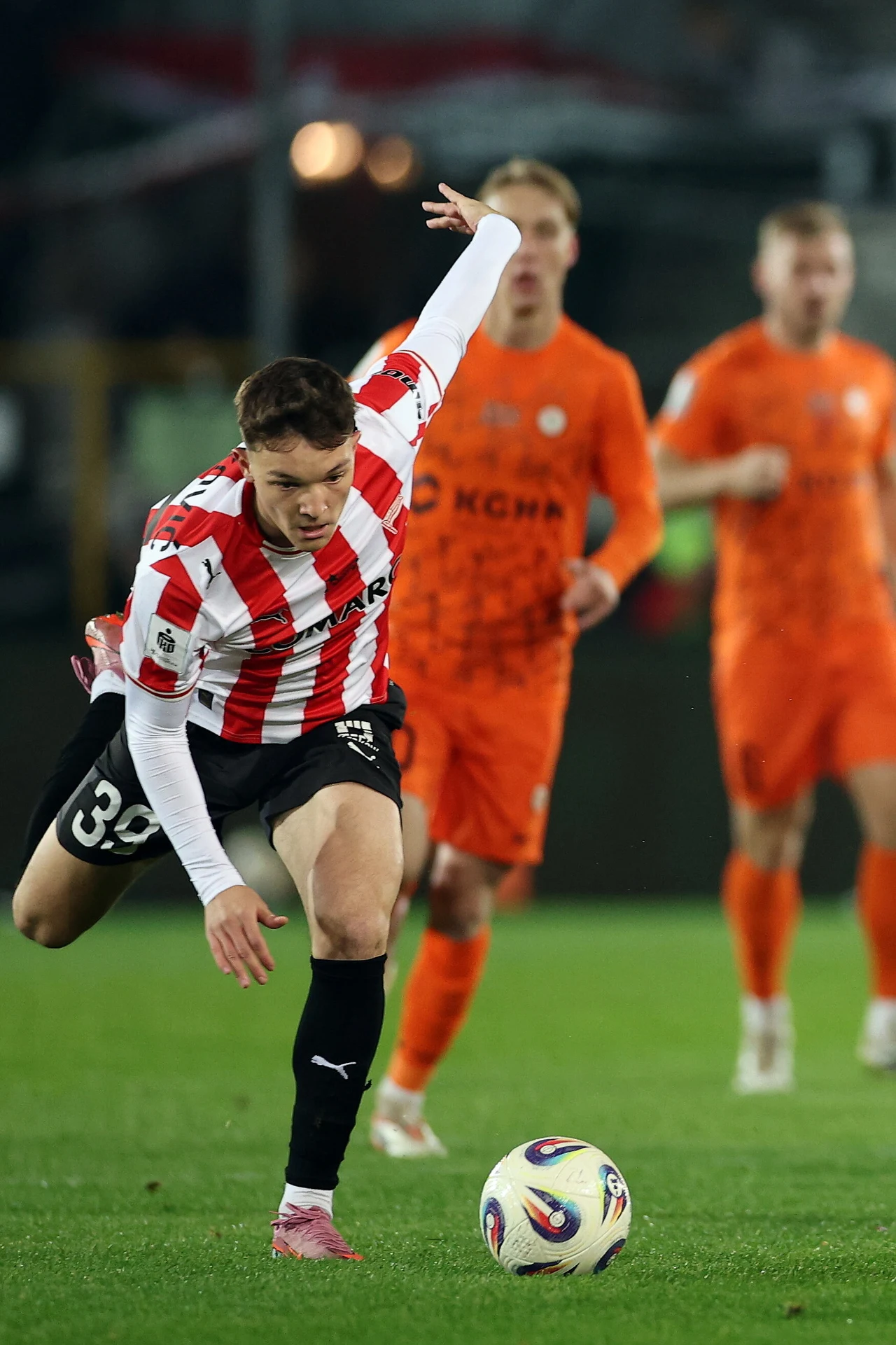 Piłkarz w biało-czerwonej koszulce walczy o piłkę podczas meczu, wyprzedzając zawodników w pomarańczowych strojach na zielonej murawie stadionu