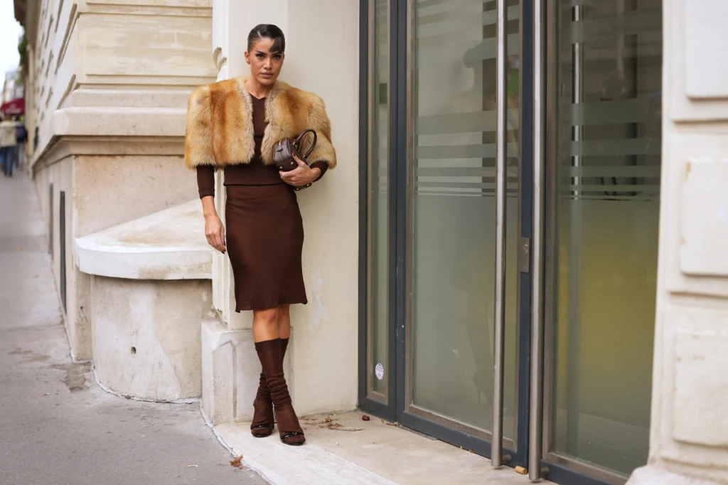 Kobieta w eleganckim brązowym stroju i futrze, trzyma kopertówkę, stoi przy jasnym budynku z przeszklonym wejściem, całość utrzymana w miejskim stylu.