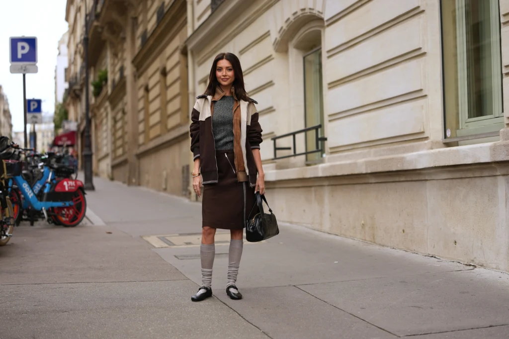 Kobieta ubrana w stylowy strój jesienny stoi na chodniku przy eleganckiej kamienicy. W tle widoczne są rowery miejskie, zaparkowane na ulicy, oraz fragmenty miejskiej zabudowy.