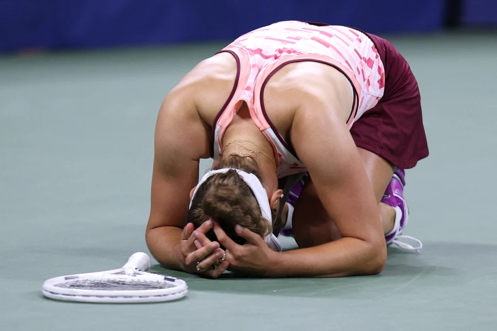Elise Mertens bierze udział w WTA Finals 2025 Zawodniczka tenisowa ubrana w różowo-białą sportową sukienkę klęczy na korcie z głową opartą na dłoniach, obok leży rakieta tenisowa. Wygląda na moment silnych emocji, prawdopodobnie wzruszenia lub wyczerpania po zakończonym meczu.
