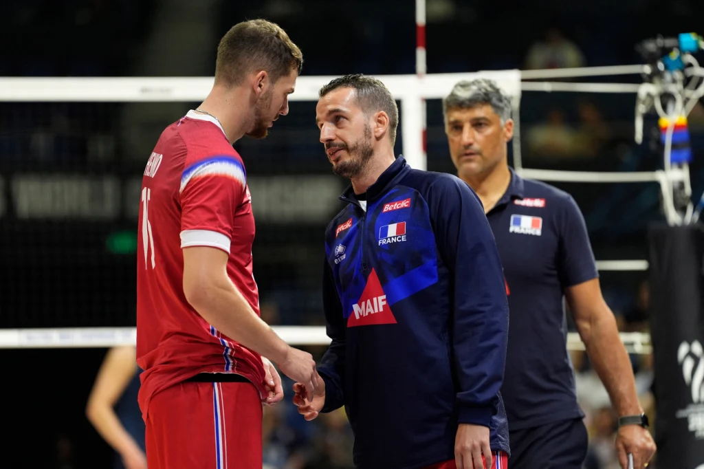 Dwóch mężczyzn znajduje się na pierwszym planie i wymienia uścisk dłoni lub rozmowę podczas meczu siatkówki, ubrani w stroje sportowe reprezentacji Francji, w tle widoczny jeszcze jeden członek sztabu oraz siatka.