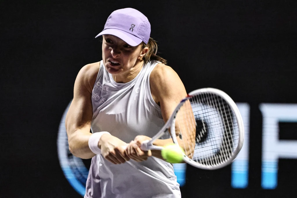 Iga Świątek w trakcie turnieju WTA Finals 2025 Zawodniczka w białym stroju sportowym uderza piłkę tenisową rakietą, skoncentrowana na grze, w tle ciemne boisko oraz rozmazane napisy.