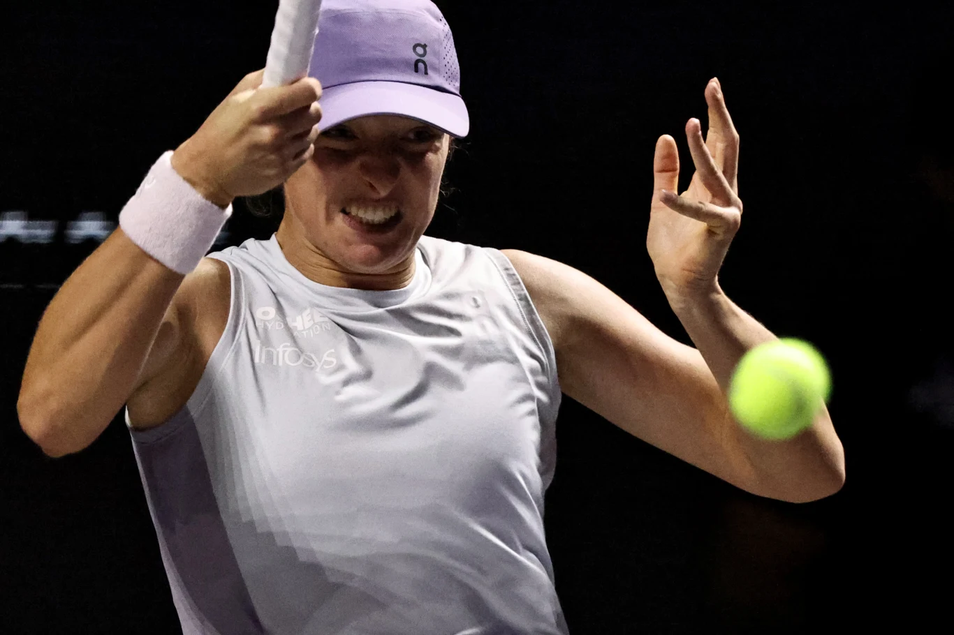 Zawodniczka w fioletowej koszulce i czapce uderzająca rakietą piłkę tenisową na czarnym tle, wyrażając intensywną koncentrację i wysiłek.