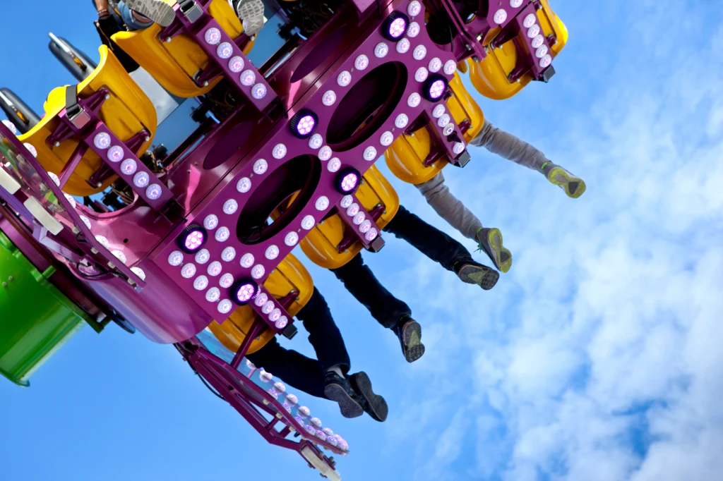 Jasnoniebieskie niebo w tle, fragment karuzeli z żółtymi siedzeniami i wystającymi nogami ludzi trzymających się za uchwyty, konstrukcja ozdobiona różowymi światłami.