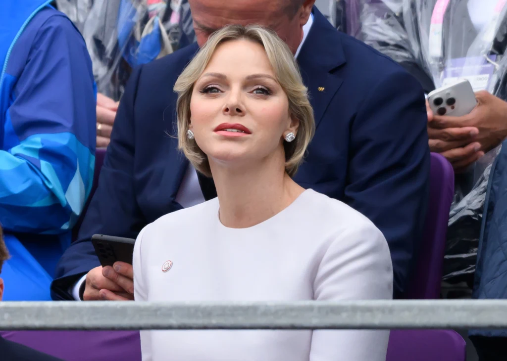 Dobra forma księżnej Charlene cieszy jej fanów i poddanych Kobieta o jasnych włosach w eleganckiej, białej kreacji siedzi wśród innych osób podczas oficjalnego wydarzenia lub ceremonii. W tle widoczne osoby w schludnych ubraniach i z telefonami komórkowymi.