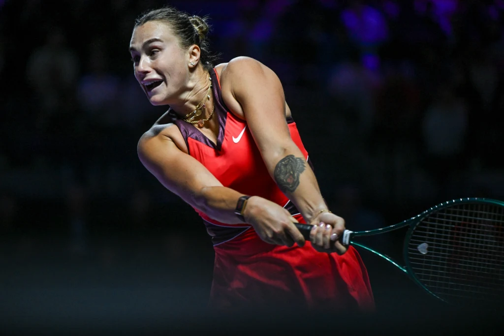 Aryna Sabalenka w trakcie WTA Finals 2025 Tenisistka w czerwonej sukience koncentruje się na odbiciu piłki rakietą podczas intensywnej gry na korcie, na tle rozmytego ciemnego tła.