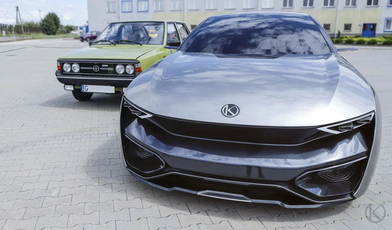 Nowoczesny srebrny samochód koncepcyjny na pierwszym planie, w tle klasyczny zielony samochód, oba zaparkowane na placu przed współczesnym budynkiem mieszkalnym. Dominują kontrasty stylistyczne między retro a futurystycznym wzornictwem aut.